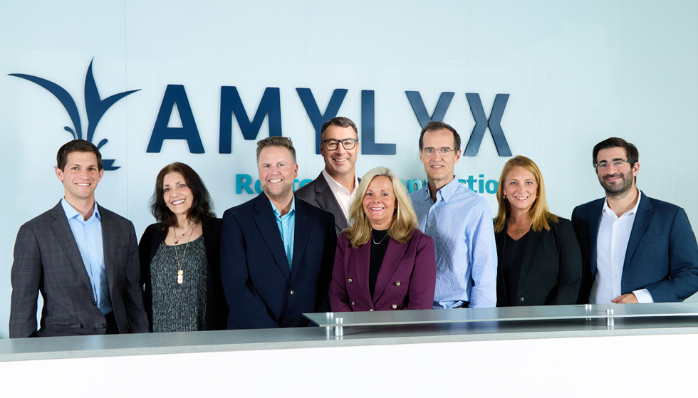 Amylyx leadership team smiling in front of the Amylyx sign. The sign reads, "Rooted in Connection."