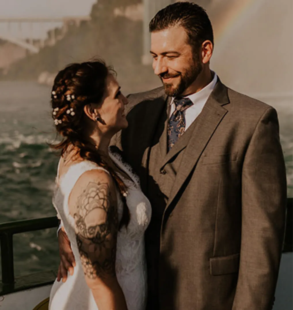 A newly married couple smiling at each other, a rainbow visible behind them 