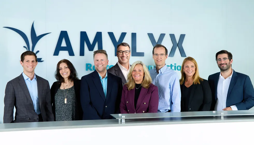 Amylyx leadership team smiling in front of the Amylyx sign. The sign reads, "Rooted in Connection."